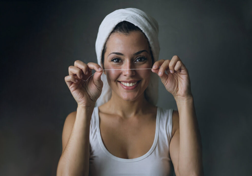 Mujer con una sonrisa sana y natural sosteniendo un aplicador de hilo dental, representando la importancia de la higiene bucal diaria.